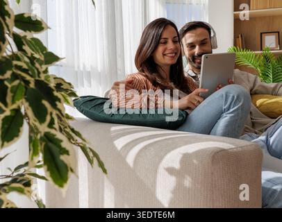 A couple sits together on a couch, smiling and looking at a tablet. They appear relaxed and engaged in listening music in a bright room. Stock Photo