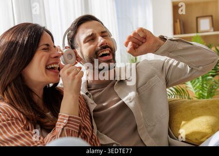Two people laugh and sing while listening to music at home. They are sitting on a couch and having a fun time. Stock Photo