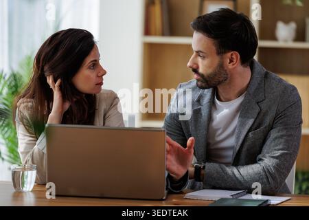 Two colleagues are discussing work while sitting at a desk. A laptop is open in front of them. They appear engaged in conversation. Stock Photo