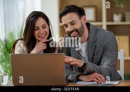 Two colleagues are discussing work while sitting at a desk. A laptop is open in front of them. They appear engaged in conversation. Stock Photo
