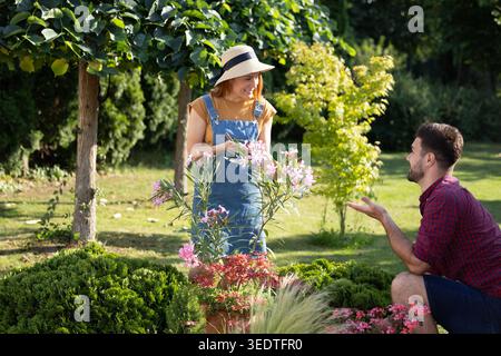 Young woman in sunhat using shears to prune flowers in garden, smiling as man requests to help while crouching Stock Photo