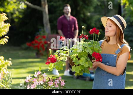 Beautiful gardener in a sunny backyard smiling while smelling bright red flowers in her hands Stock Photo
