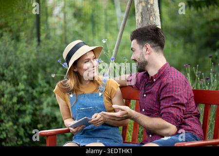 Attractive couple sitting on a bench in backyard, woman has a tablet in hand while happily looking at man while chatting Stock Photo