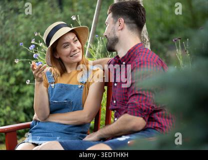 Young man and woman sit on a bench in a garden. They are smiling and enjoying a conversation surrounded by plants and flowers. Stock Photo