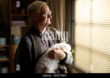 Senior Man Holding Pet Dog and Looking through Window at Home Stock Photo