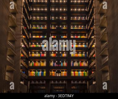 Symmetrical view of a modern building atrium at night with warm lights and colorful window panels across multiple floors. Stock Photo