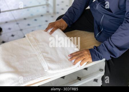 Calm person folding clean white towel by hand on wooden table completing daily laundry chore showing routine domestic housework organizing neat fabric Stock Photo