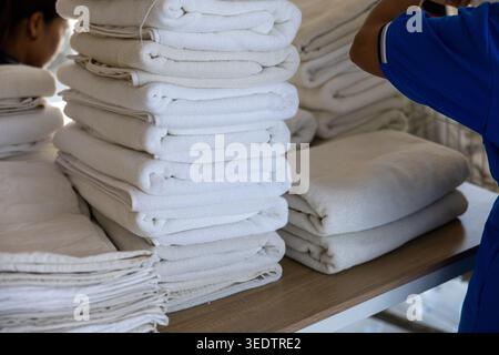 Focused laundry worker folding clean white towel stack for hotel housekeeping service working diligently behind large piles of freshly washed soft lin Stock Photo