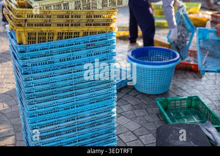 Focused worker sorting goods near tall stack of blue and yellow plastic crate container at an outdoor market storage area handling heavy equipment and Stock Photo