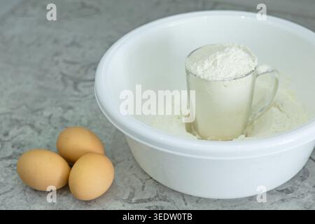 White mixing bowl with flour, glass mug filled with flour, and three brown eggs on kitchen counter. Stock Photo