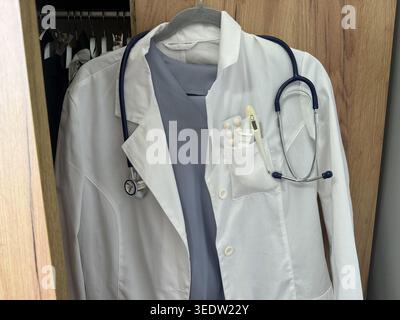 A white coat is hanging on a hanger. The coat has a stethoscope attached to it. Medical supplies can be seen in the coat pocket. The setting looks lik Stock Photo