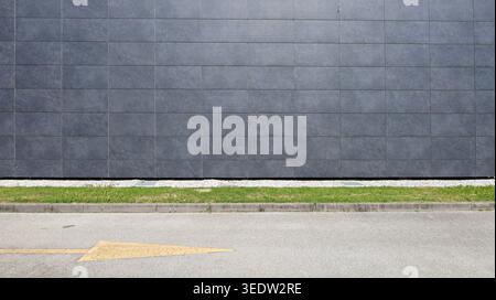 Gray stone tiles coating of a ventilated facade at roadside with grassy sidewalk and street in front. Background for copy space. Stock Photo