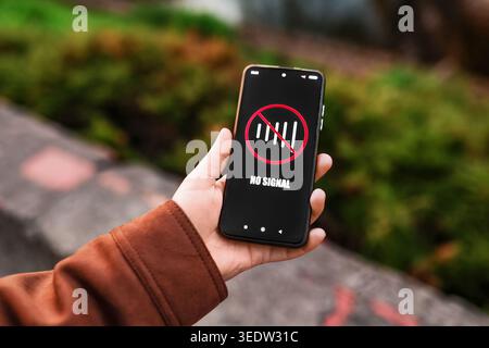Hand holding a smartphone displaying a "No Signal" icon with a crossed-out network bar outdoors. Concept of poor cellular connection, internet outage, Stock Photo