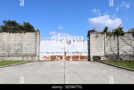 No stopping sign on a locked white and rusty gate with security spikes. Concrete surrounding wall by sides, sky above. Background for copy space Stock Photo