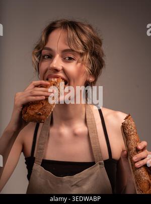 Hungry young woman in apron biting fresh crusty sourdough baguette, enjoying a delicious snack Stock Photo