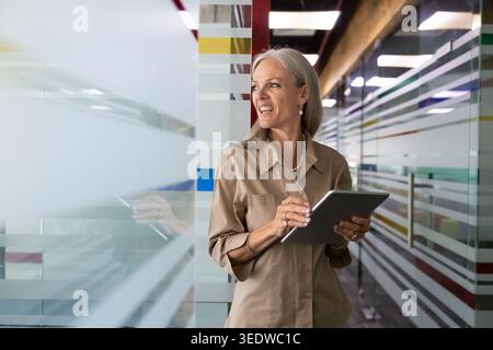 Middle age businesswoman stands in office hallway with digital tablet Stock Photo