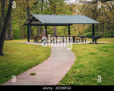 Empty outdoor picnic shelter with tables and a charcoal grill at the end of a paved path in a lush green forest preserve during the spring season Stock Photo