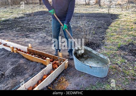 Worker Mixing Mortar For Outdoor Foundation. Worker mixes wet mortar in a metal tub beside wooden formwork and brick rubble for a small garden Stock Photo