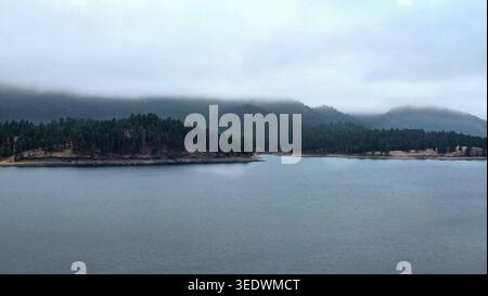 Misty Forest Lake In Mountain Landscape. Calm lake surrounded by pine-covered hills and low clouds, creating a peaceful misty mountain landscape. Stock Photo