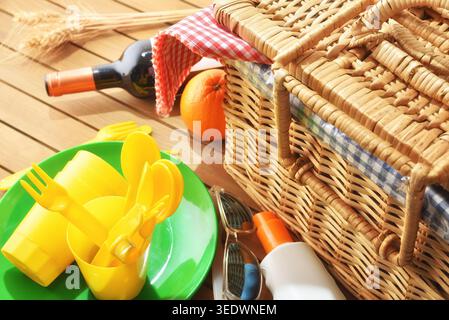 Close-up of picnic utensils, wicker basket, wine bottle, orange and sunscreen on a wooden table in bright sunlight. Stock Photo