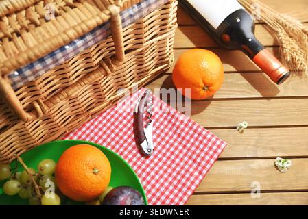 Top view of picnic setup with wicker basket, wine bottle, oranges, grapes and corkscrew on a wooden table outdoors. Stock Photo