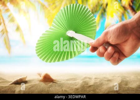Close-up of an adult hand holding a green paper fan on a sunny tropical beach with palm trees and seashells in the background. Stock Photo