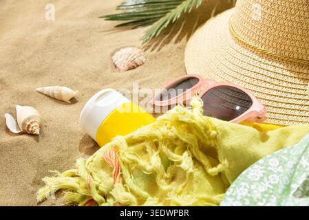 Beach accessories including straw hat, sunglasses, sunscreen, scarf, and seashells arranged on warm sand in bright sunlight. Stock Photo