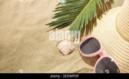 Close-up of pink sunglasses, straw hat, palm leaf, and seashell on sandy beach with bright tropical summer lighting. Stock Photo