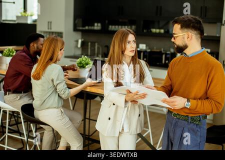 In a bright office, two people talk while holding documents. Two others work on laptops. The setting shows a busy and collaborative work environment. Stock Photo