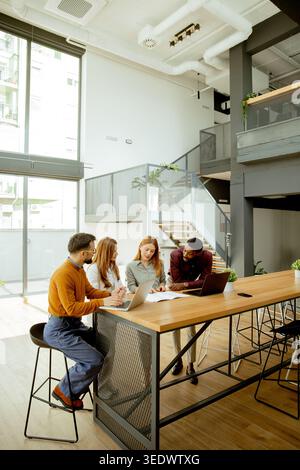 Four people are seated at a table in a modern office. They are using a laptop and discussing ideas. The space has large windows and a stairway in the Stock Photo