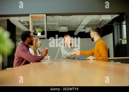 Four professionals meet in an office setting. Two people shake hands at a long table while two others observe. Laptops and drinks are placed on the ta Stock Photo