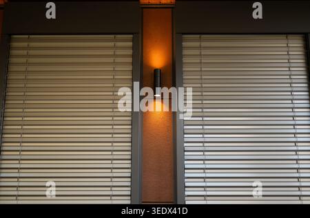 Modern building facade featuring metal external blinds and a warm vertical wall lamp between windows. Stock Photo