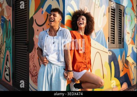 Joyful man and woman looking at each other with love while holding hands against a bright wall Stock Photo