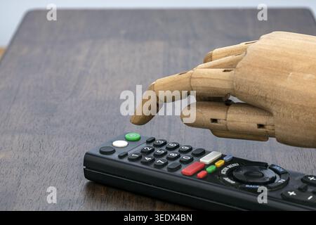 A jointed wooden artist mannequin hand hovers over a black Smart television remote control on a dark wooden table, with the index finger pointing down Stock Photo