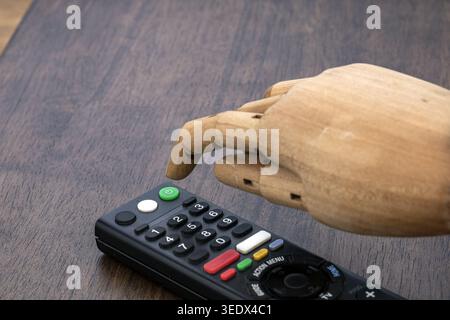 An articulated wooden artist mannequin hand extends its index finger to press the green power button on a black Smart TV remote control resting on a d Stock Photo