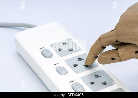 A wooden jointed mannequin hand pokes its finger directly into a live electrical outlet on a white multi-socket power strip. This striking image power Stock Photo