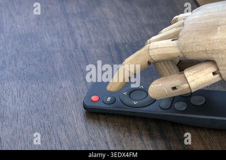 A detailed wooden articulated robot hand with jointed fingers precisely presses a navigation button on a sleek black remote control, resting on a dark Stock Photo