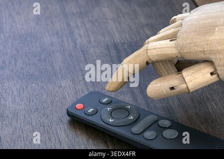 An articulated wooden robot hand with jointed fingers points its index finger at a black TV remote control featuring ON, OFF, and navigation buttons o Stock Photo