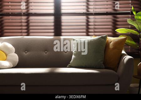 Colorful pillows on soft sofa in front of blinds indoors Stock Photo