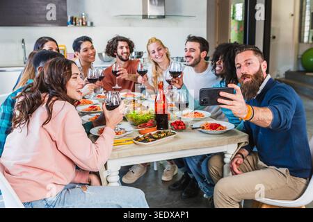 Diverse friends enjoying dinner, toasting with wine, and taking a selfie at home Stock Photo