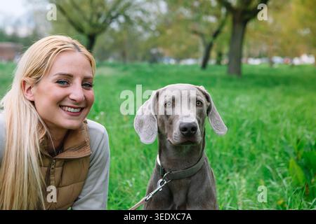 Young woman and her weimaraner sitting together in green grass, smiling Stock Photo