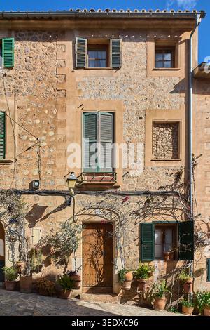 Traditional stone house facade with green shutters and potted plants on a narrow street in a Mediterranean village during a sunny day with shadows Stock Photo