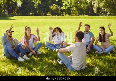 Group of young multiethnic adults sitting in circle on grass, raising hands, engaged participating Stock Photo