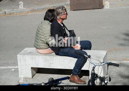 A unidentified couple sits back-to-back resting on a concrete ledge along an open street space on a bright, sunny day, Girona, Spain. Stock Photo