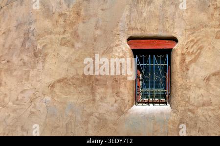 Small, narrow window on adobe home has a red mantel.  Metal grate over window is ornate and weathered blue. Stock Photo