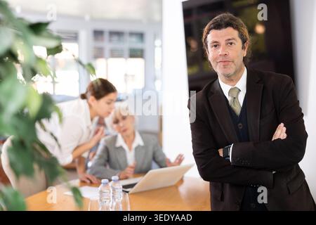 positive male colleague standing in meeting room and employees are talking behind him at table Stock Photo