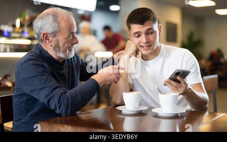 Meeting at cafe. Guy person scrolls down phone screen, elderly man comforts his son Stock Photo