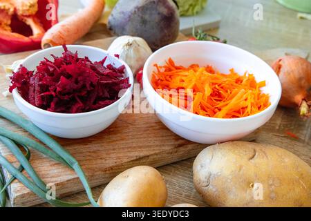 Shredded beets and carrots sit in white bowls on a wooden board. Nearby, potatoes, onions, and other vegetables are arranged for prepping a meal. The Stock Photo