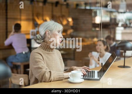 Positive European old woman drinking and enjoying coffee while working on laptop in cafe Stock Photo