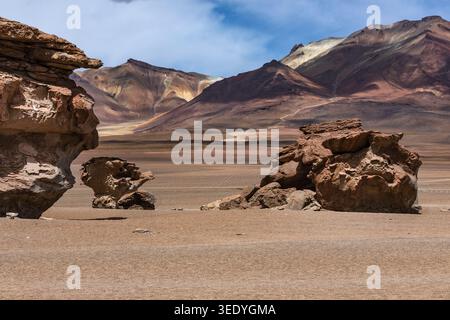 Wind-eroded rock formations rise from arid desert floor in front of colorful Andean mountains, Siloli Desert, Bolivian Altiplano, Bolivia Stock Photo